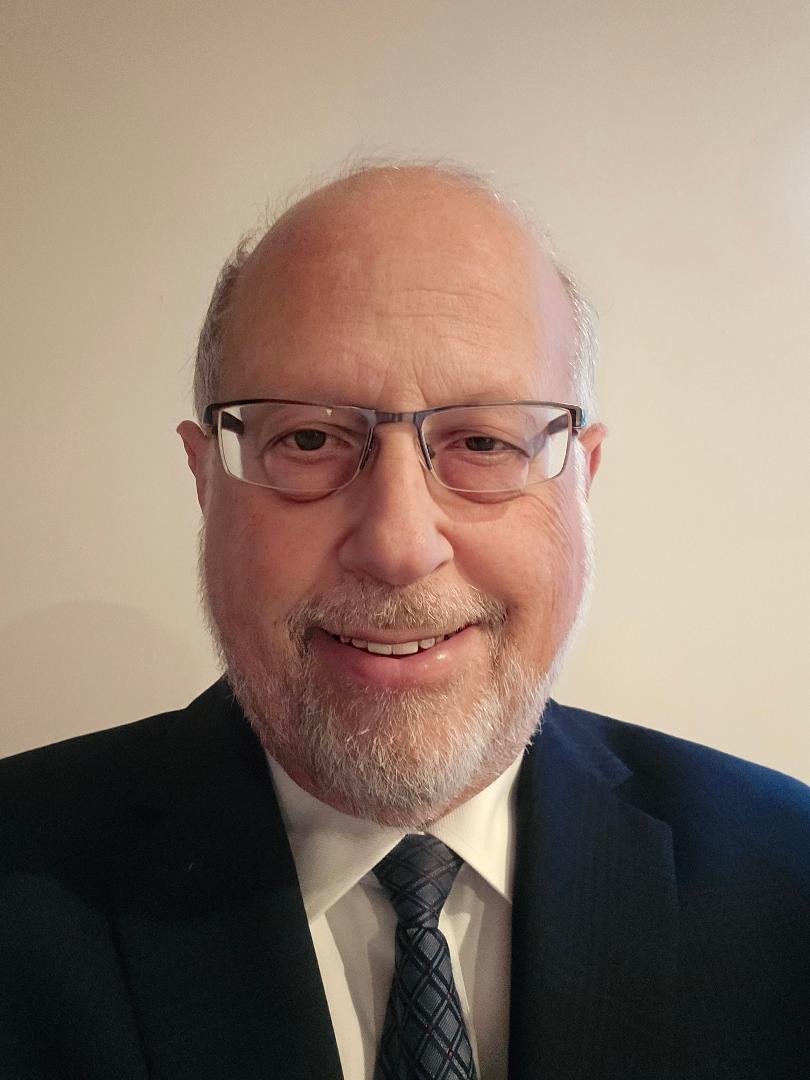 The image shows a man wearing glasses, smiling at the camera, with gray hair and a beard. He is dressed in a suit and tie.