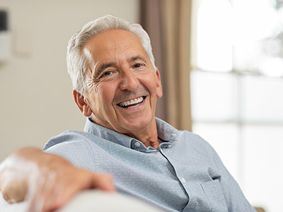 The image shows an older man with gray hair smiling at the camera while seated on a couch indoors.