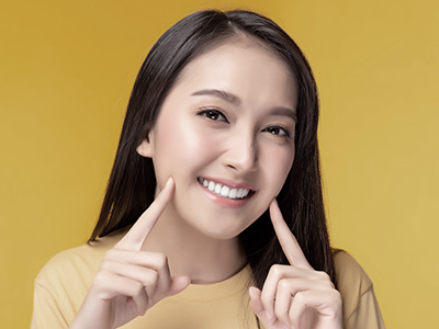 A woman with light skin smiles at the camera while making a peace sign gesture with her hand. She has long hair, wears a yellow top, and stands against a solid mustard background.