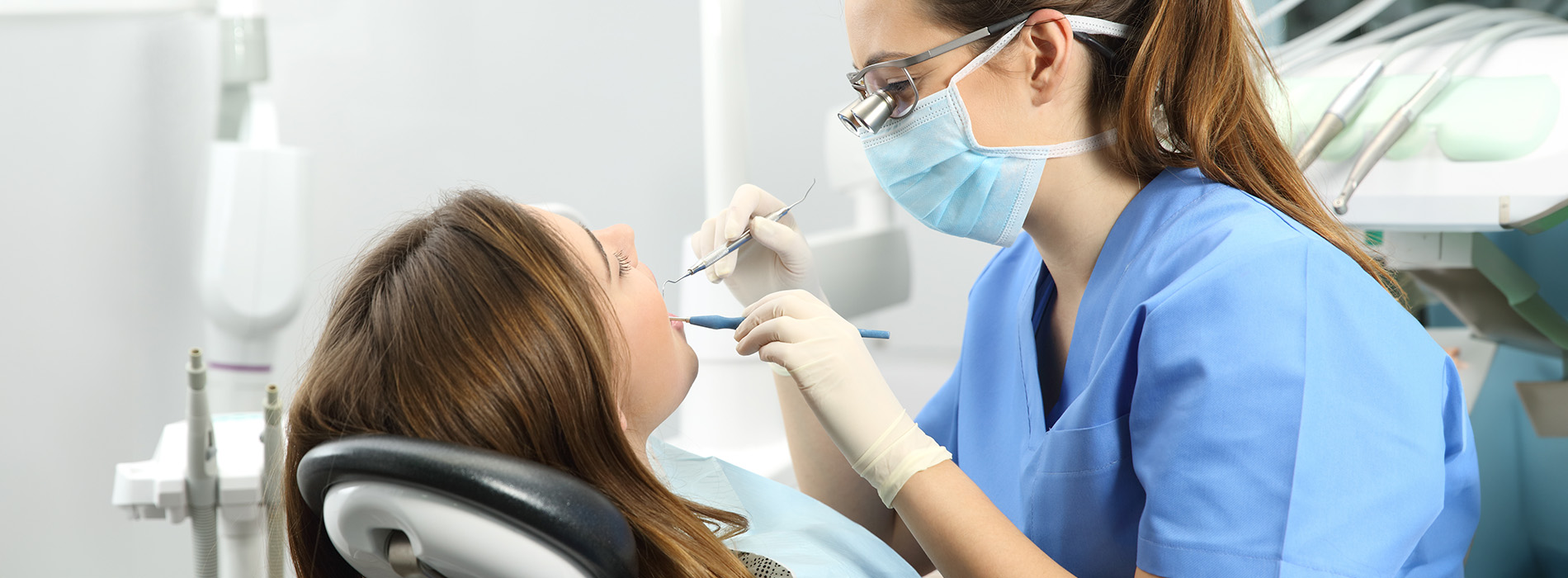 A dental hygienist in a blue scrub uniform is performing a teeth cleaning procedure on a patient s mouth using dental tools, with the patient seated in an adjustable dental chair.