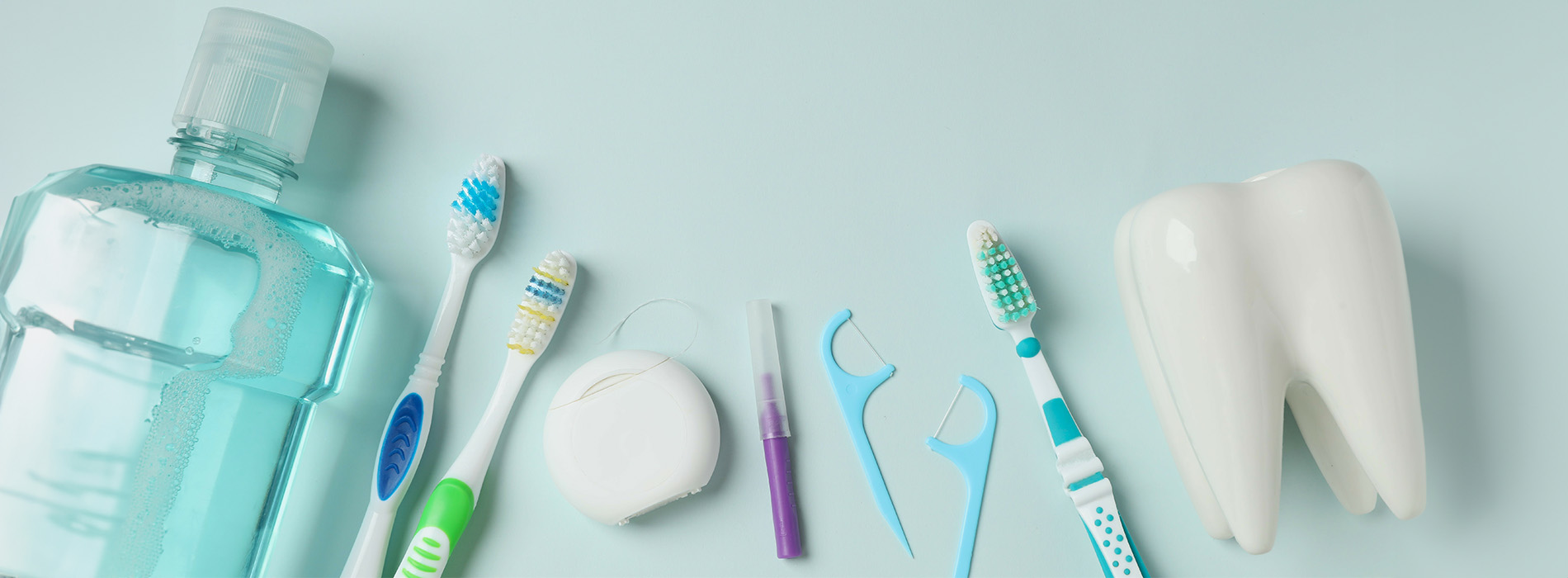 The image displays a collection of dental hygiene items including toothbrushes, toothpaste, mouthwash, floss, and dental picks arranged on a light background with a water bottle and other personal care products in the foreground.