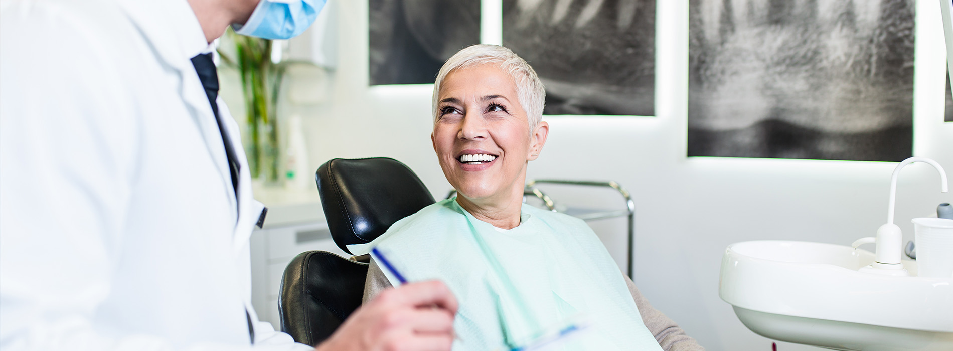 A woman seated in a dental chair with a smiling expression, receiving attention from a standing dentist who is holding a pen, set against a backdrop of a dental office interior with various equipment and tools visible.