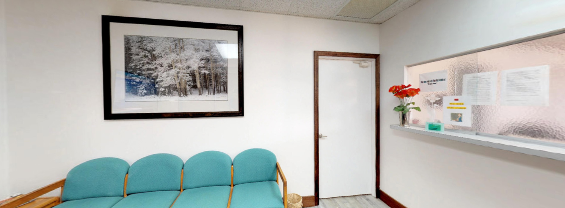 The image shows an interior space with a waiting area featuring a blue sofa, a framed picture on the wall, and a door leading to another room. There is a window with curtains partially drawn, allowing natural light into the space.