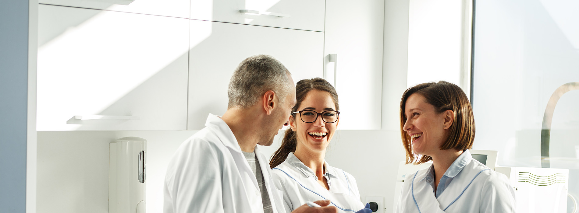 The image shows three individuals in a professional setting, likely a dental office, with one person seated at a counter and two standing behind, engaging in conversation.