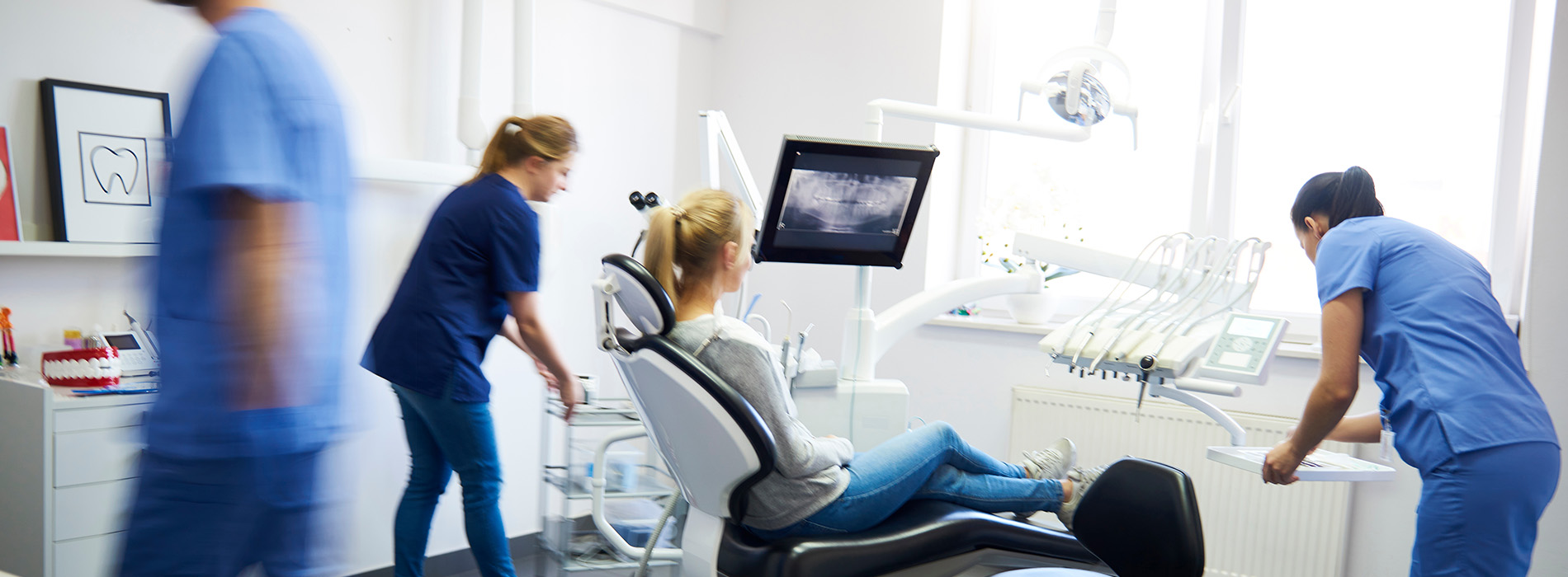 The image shows a group of healthcare professionals working together in a medical setting, with a patient being attended to by a nurse and a doctor using technology such as a monitor and a computer screen.