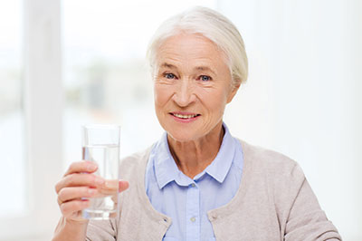 The image shows an elderly woman holding a glass of water with her right hand while standing indoors, wearing a light blue cardigan and smiling at the camera.