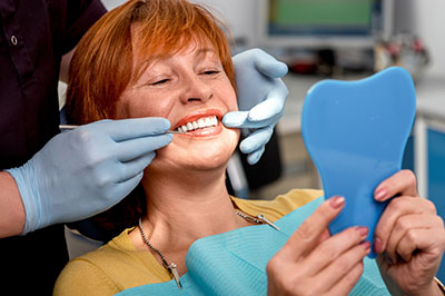 In the image, a woman with red hair is sitting in a dental chair, holding a blue toothbrush, while a dentist wearing gloves prepares her teeth for cleaning.