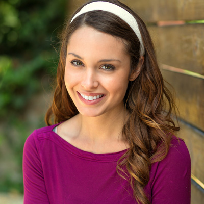 A woman with long hair wearing a purple top poses against a wooden fence background.