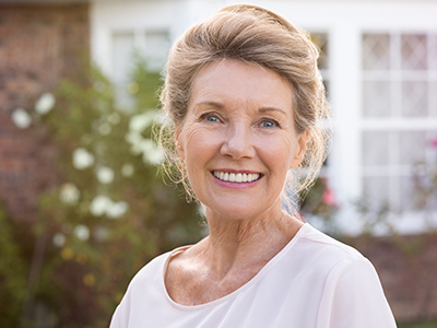 An elderly woman wearing a light-colored top stands outdoors with a house in the background.