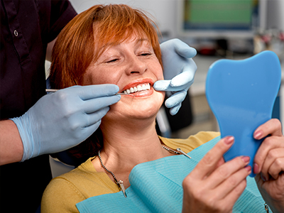 A woman with red hair sitting in a dental chair, smiling at the camera while a dentist works on her teeth, holding a blue mouthguard.