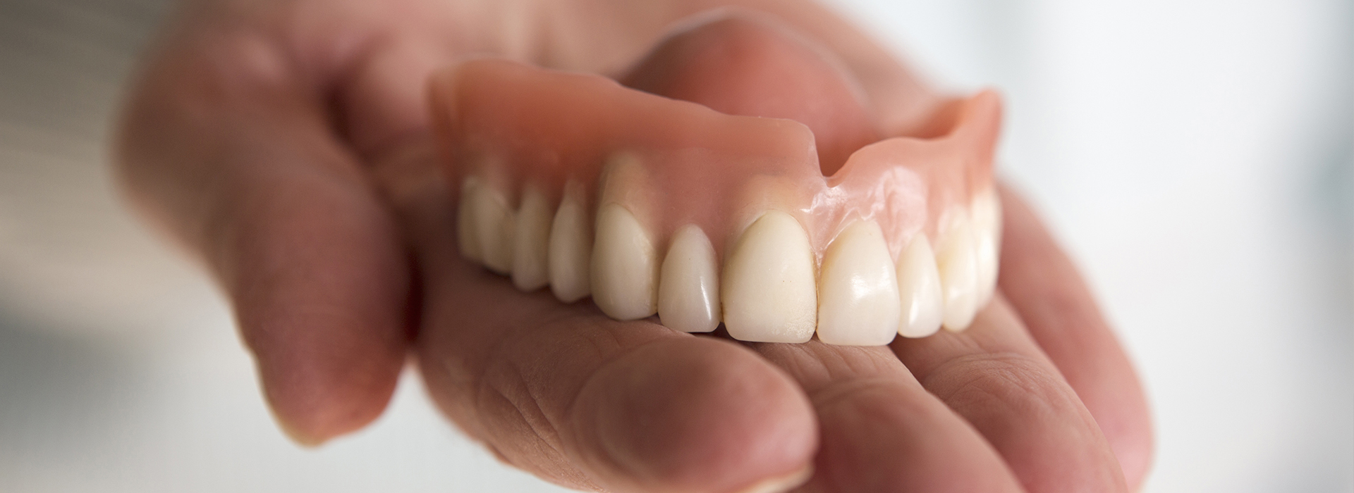 The image shows a person holding an artificial set of teeth with a neutral background.