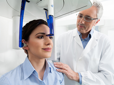 The image depicts a medical setting where a woman is seated in front of a large scanner machine with a blue headpiece, while a male doctor stands beside her, engaging in conversation.