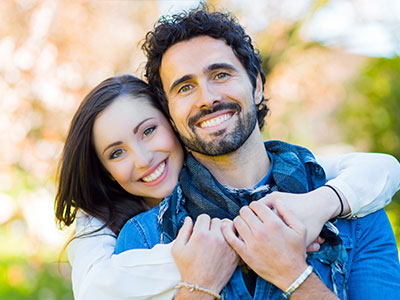 The image shows a man and woman hugging each other closely, both smiling, with the man wearing a scarf around his neck, standing outdoors during daylight.