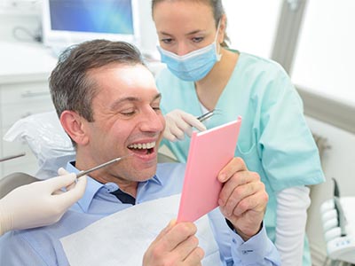 A man sitting in a dental chair with a pink cardboard sign held up in front of him, smiling at the camera while a woman in scrubs looks on, holding dental tools.