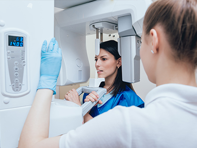 A woman in a blue uniform stands beside a large white 3D scanner machine, with another person partially visible behind her, both in an indoor setting.