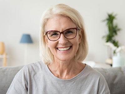 The image features a smiling middle-aged woman with short blonde hair, wearing glasses, a gray top, and sitting on a couch. She has a warm expression and appears to be indoors, possibly in a living room setting.