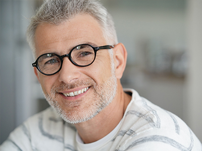 The image shows a man with glasses, smiling at the camera, against a blurred background that appears to be an indoor setting.