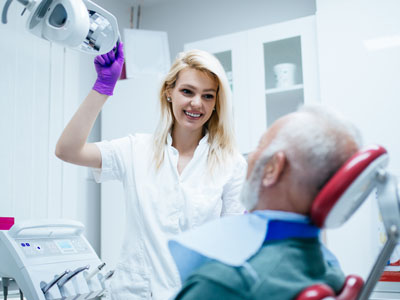 A woman in a white lab coat stands by a man in a dental chair, holding a tray with dental instruments, both are in a dental office setting.