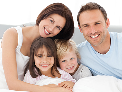 A family of four, including two adults and two children, smiling together on a bed with a white comforter.