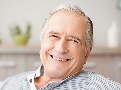 The image shows an elderly man with gray hair smiling at the camera while seated comfortably indoors.