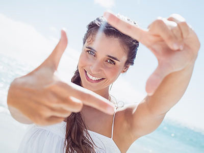 A woman with long hair is smiling at the camera while holding up two fingers, creating a peace sign gesture against a bright blue sky background.